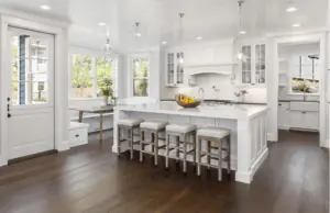 A modern kitchen with white cabinetry, a large center island with stools, and dark wooden flooring. The kitchen features a farmhouse sink, stainless steel appliances, and a window seat. Large windows bring in natural light, and pendant lights hang above the island.