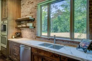 Modern kitchen with a marble countertop, brick backsplash, wooden cabinets, open shelving, and a large window showcasing a scenic outdoor view.